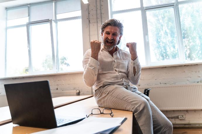 Hombre feliz sentado sobre la mesa en pose de victoria mirando la pantalla de un ordenador