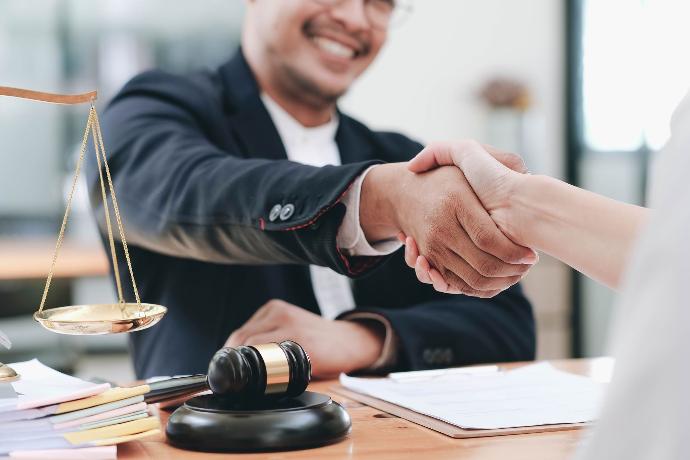 hombre en traje alegre estrechando manos con una cliente, en su mesa hay un papel junto a un mazo y a una balanza